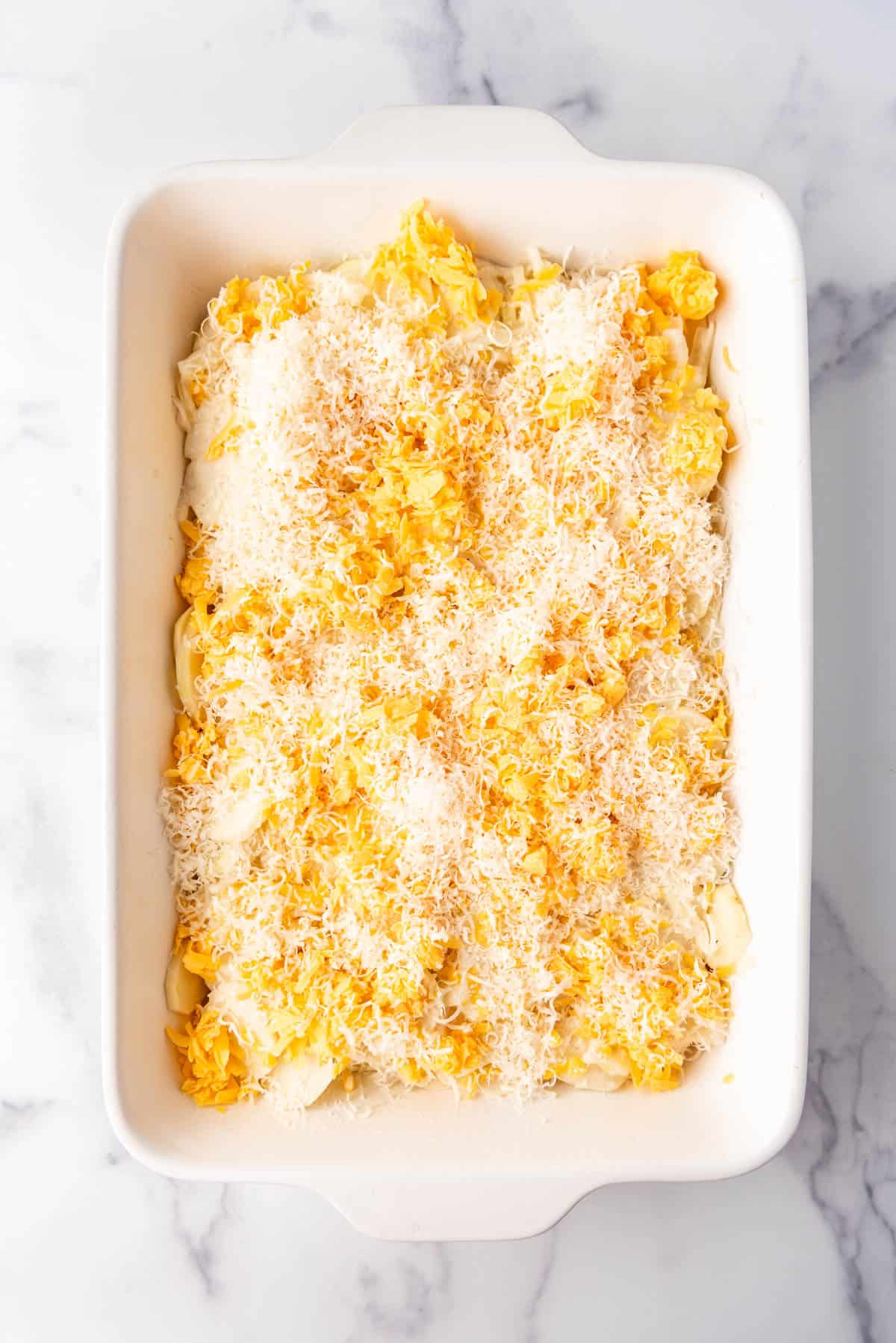 Grated parmesan and cheddar cheeses sprinkled over potatoes in a white casserole dish.