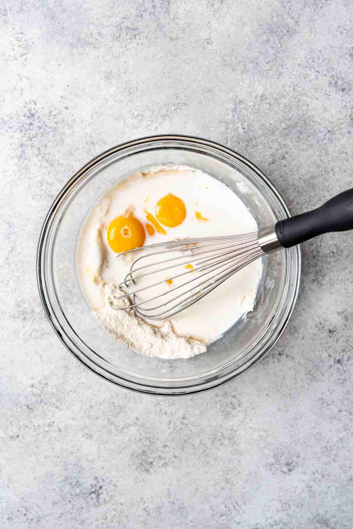 An image of flour, sugar, egg yolks, and milk in a bowl with a whisk.