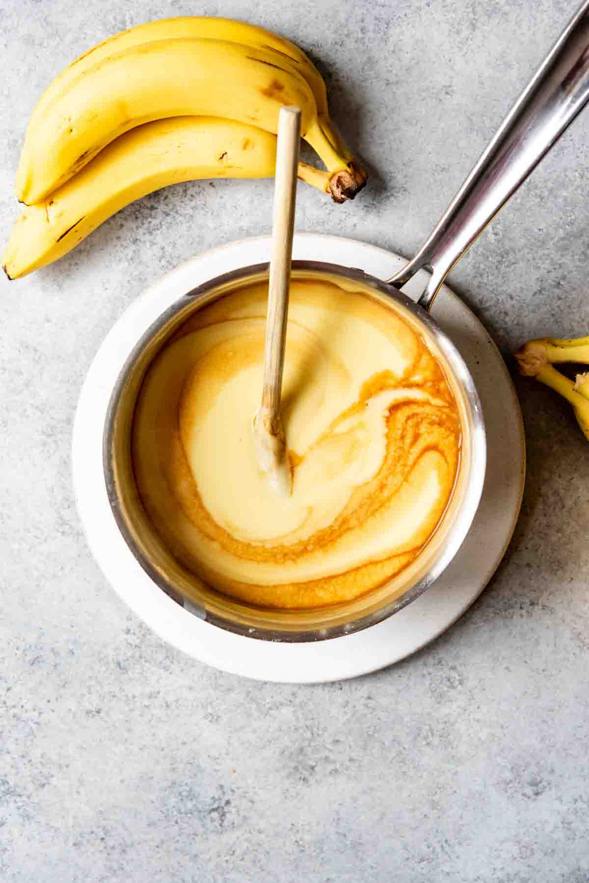 An image of homemade vanilla pudding in a pan with vanilla being swirled into it and bananas off to the side.