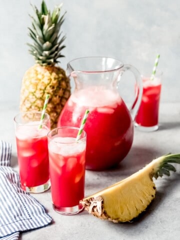 An image of a pitcher of homemade Hawaiian punch with glasses of punch served on ice beside it.
