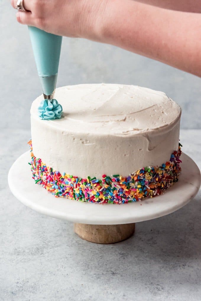 An image of a hand holding a piping bag, piping swirls onto a frosted funfetti cake.