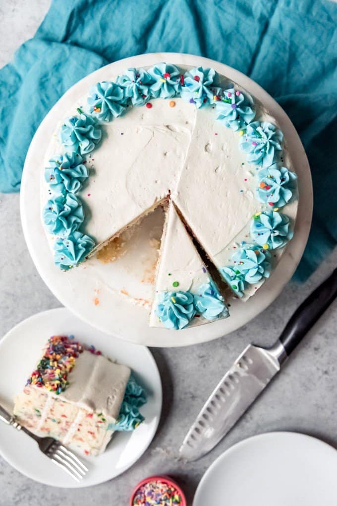 An image of a partially sliced funfetti cake with a slice sitting on a white plate next to the cake.