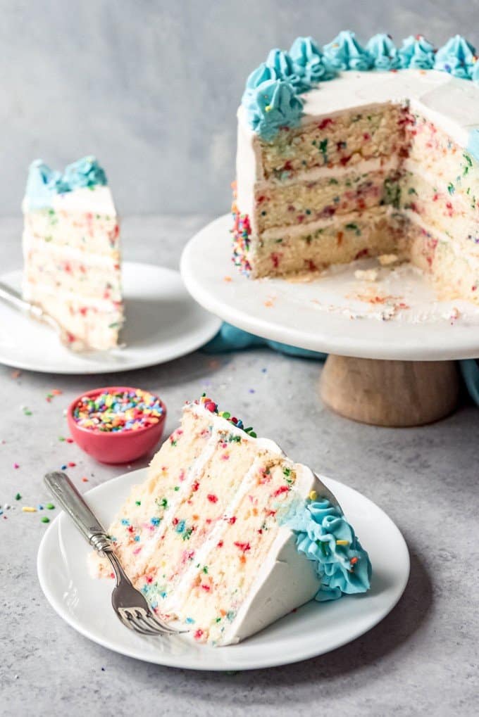 An image of a slice of funfetti cake on a plate with a fork next to the sliced cake on a cake stand.