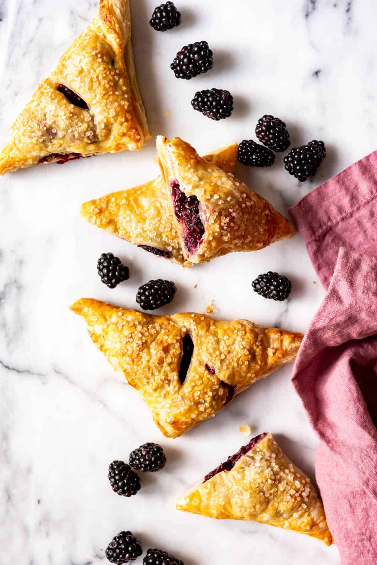 An image of triangle-shaped blackberry turnovers on a marble slab with fresh blackberries scattered around.