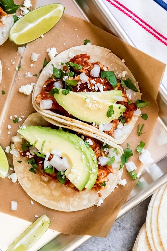 An image of two shredded chicken tinga tacos topped with avocado slices, cotija cheese, chopped onions, cilantro, and a squeeze of lime juice.