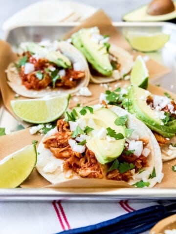 An image of smoky shredded chicken tinga tacos topped with sliced avocado, cilantro, chopped onion, cotija cheese, and lime juice.