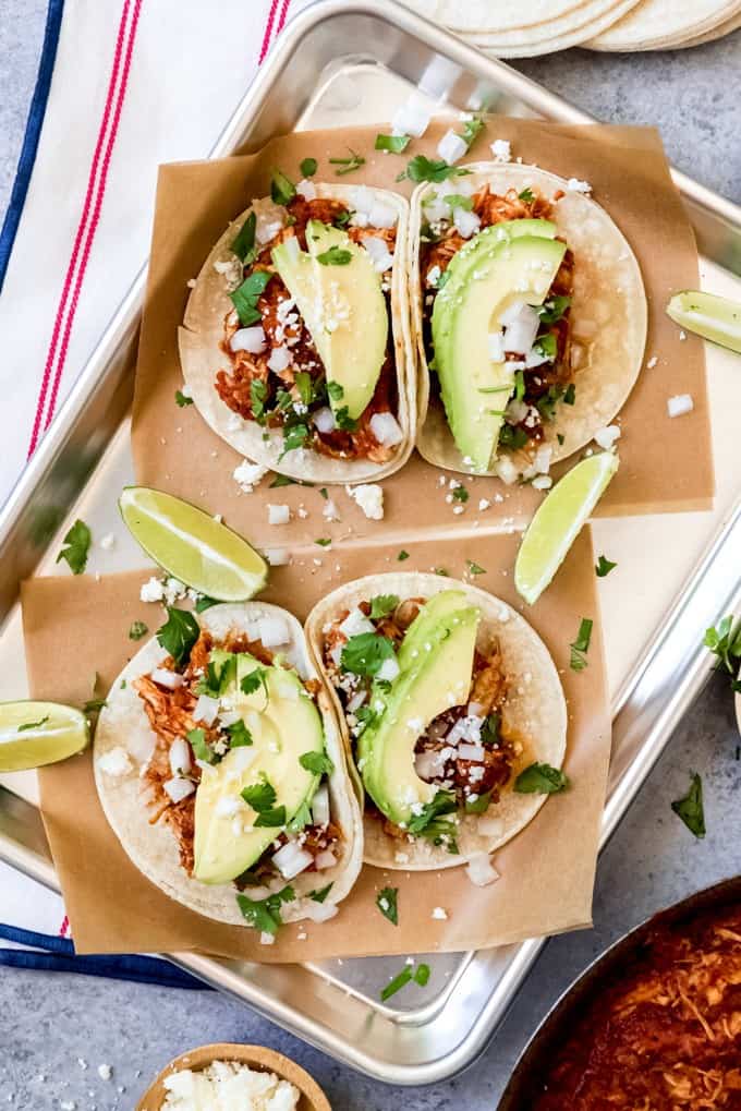An image of smoky chicken tinga tacos topped with sliced avocado, cotija cheese, chopped onion, cilantro, and lime juice.