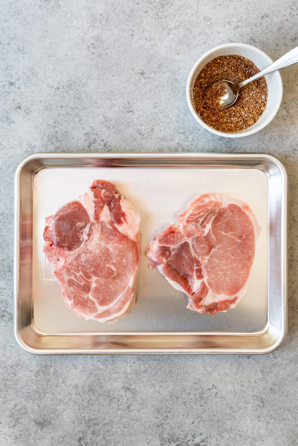 An image of two bone-in pork loin chops on a tray next to a bowl of homemade spice rub.