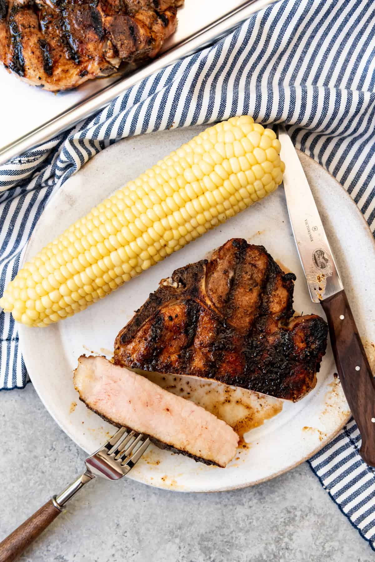 An image of a slice of grilled pork chop with a blush of pink on the inside on a plate with boiled corn on the cob.