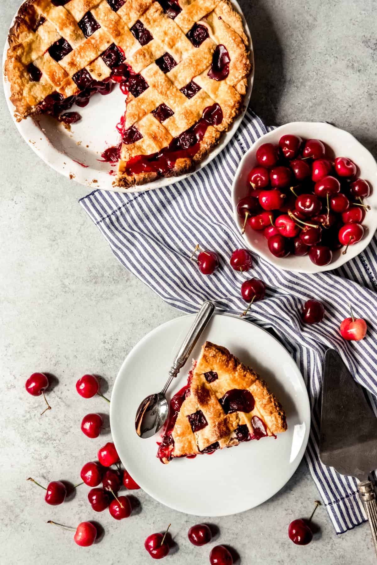An image of a slice of classic cherry pie with lattice crust on a plate next to the whole pie and a bowl of fresh cherries.
