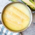 An image of ears of corn on the cob being boiled in a large pot with milk and butter.