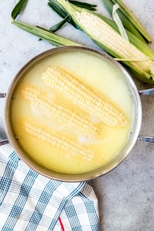 An image of ears of corn on the cob being boiled in a large pot with milk and butter.