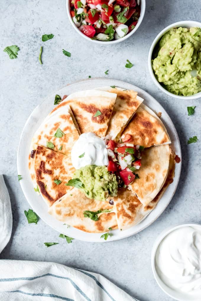 An image of quesadillas piled on a plate with sour cream, guacamole, and pico de gallo on top.