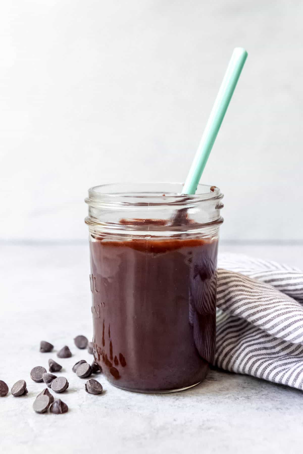 A jar of homemade hot fudge with chocolate chips and a striped linen napkin beside it.