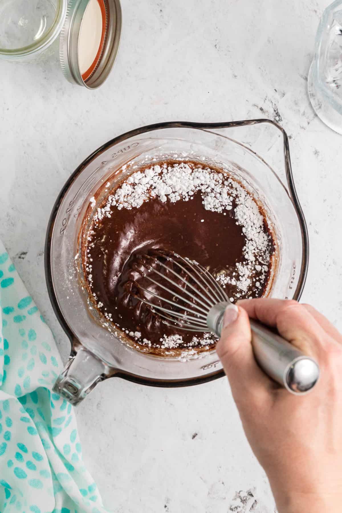 A hand holding a whisk whisking ingredients to make hot fudge sauce.