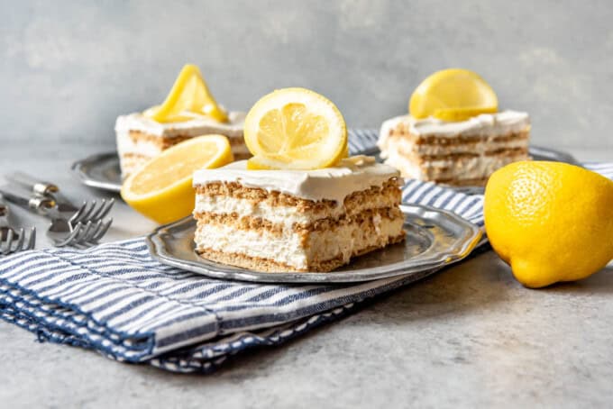 Three slices of lemon icebox cake on plates next to lemon halves.