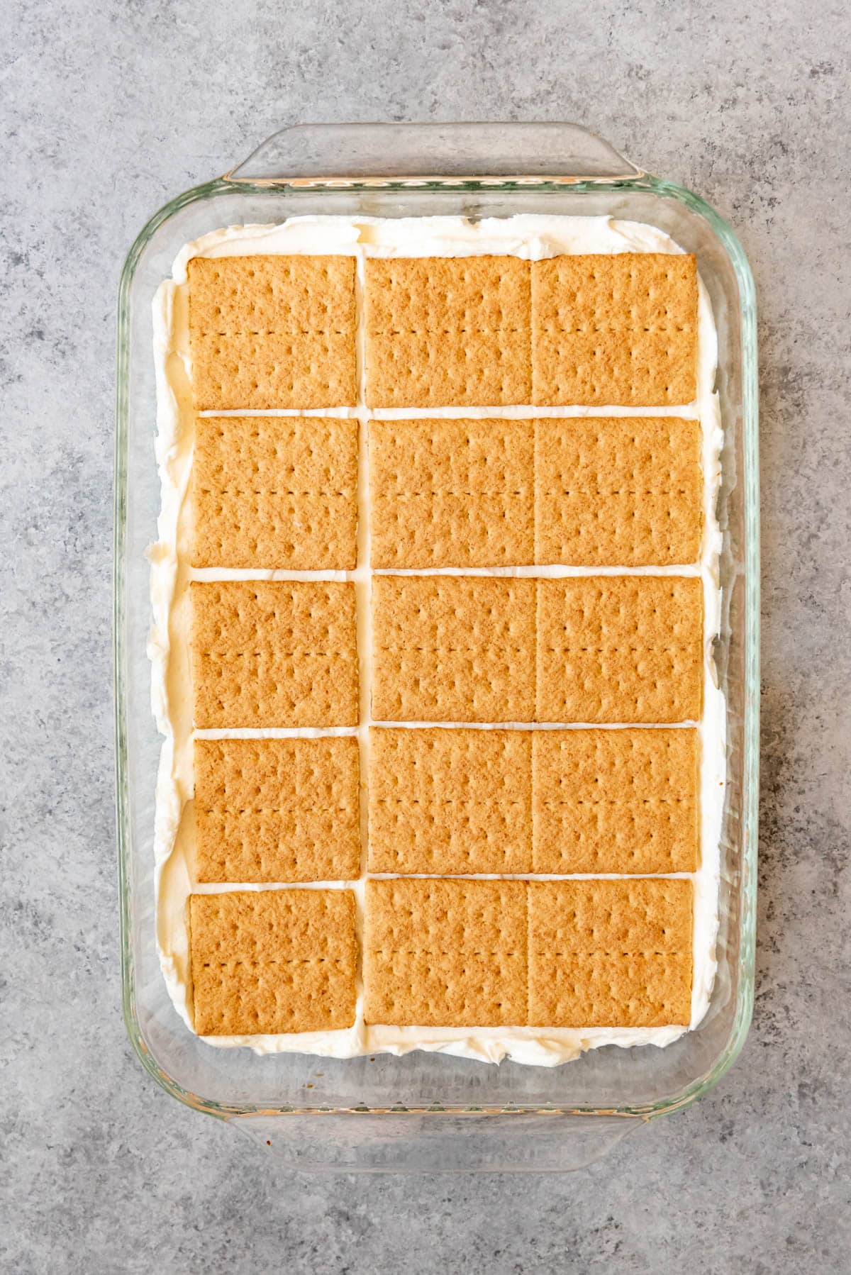 An image of graham cracker squares arranged on top of a creamy lemon layer for a no-bake lemon icebox cake.