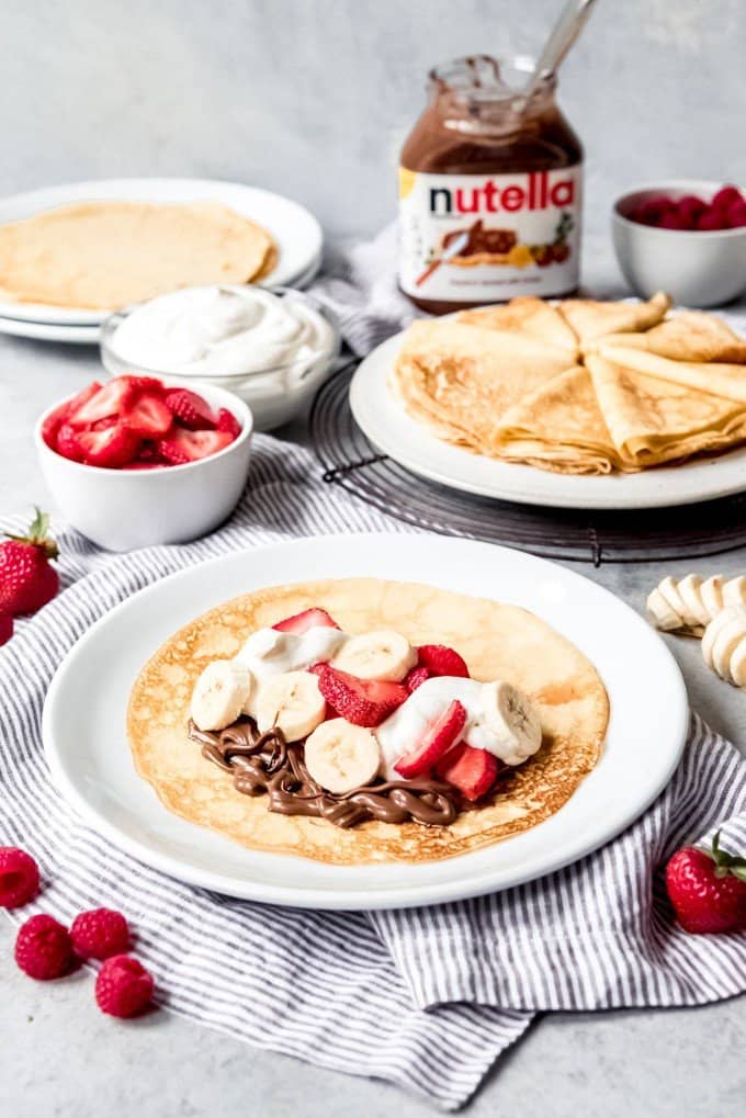 An image of stacks of warm sweet crepes with toppings like nutella, berries, and cream, with one already-filled crepe on a plate.