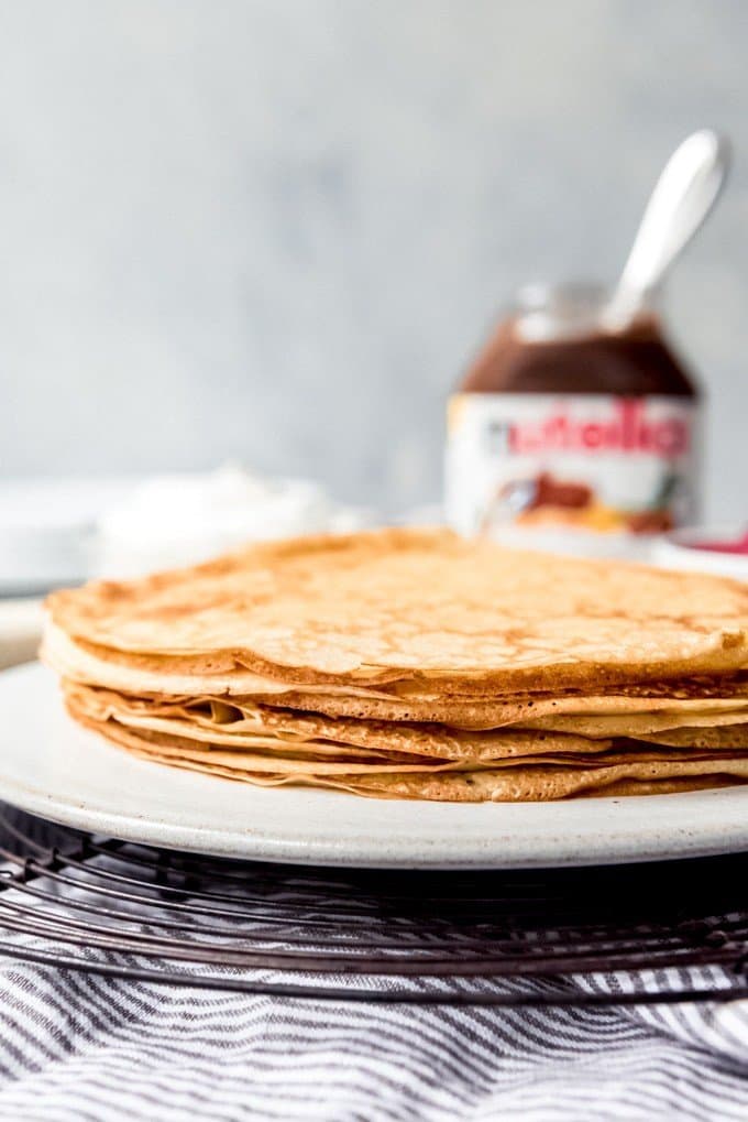 An image of a stack of thin, golden brown crepes on a plate with a jar of nutella behind them.