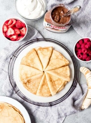 An image of light and tender homemade crepes stacked on a plate with nutella, fruit, and whipped cream for filling.