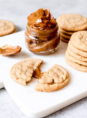 An image of a brown sugar cookie stuffed with dulce de leche with a jar of dulce de leche behind it.