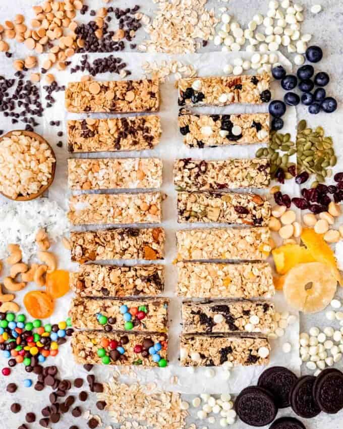 An image of eight different flavors of homemade granola bars arranged on a surface with ingredients spilling around them.