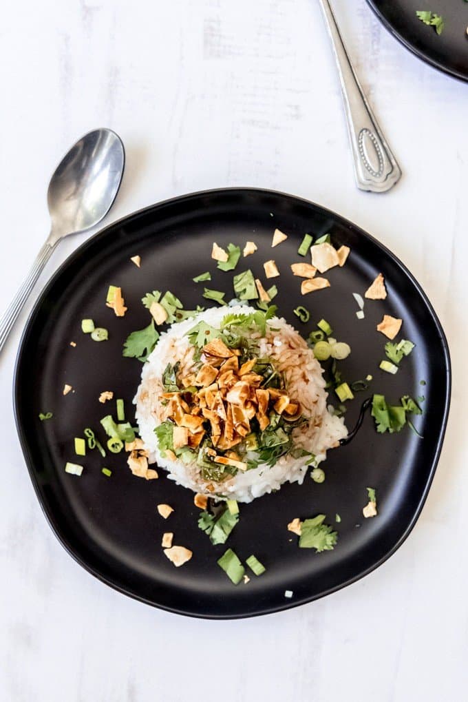 An image of a black plate with a mound of glutinous rice topped with cilantro, soy glaze, and crispy garlic chips.