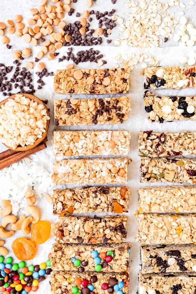 An image of different flavors of homemade chewy granola bars with lots of mix-ins around them like peanut butter chips, mini chocolate chips, rice krispies, coconut, cashews, dried apricots, and M&M's.