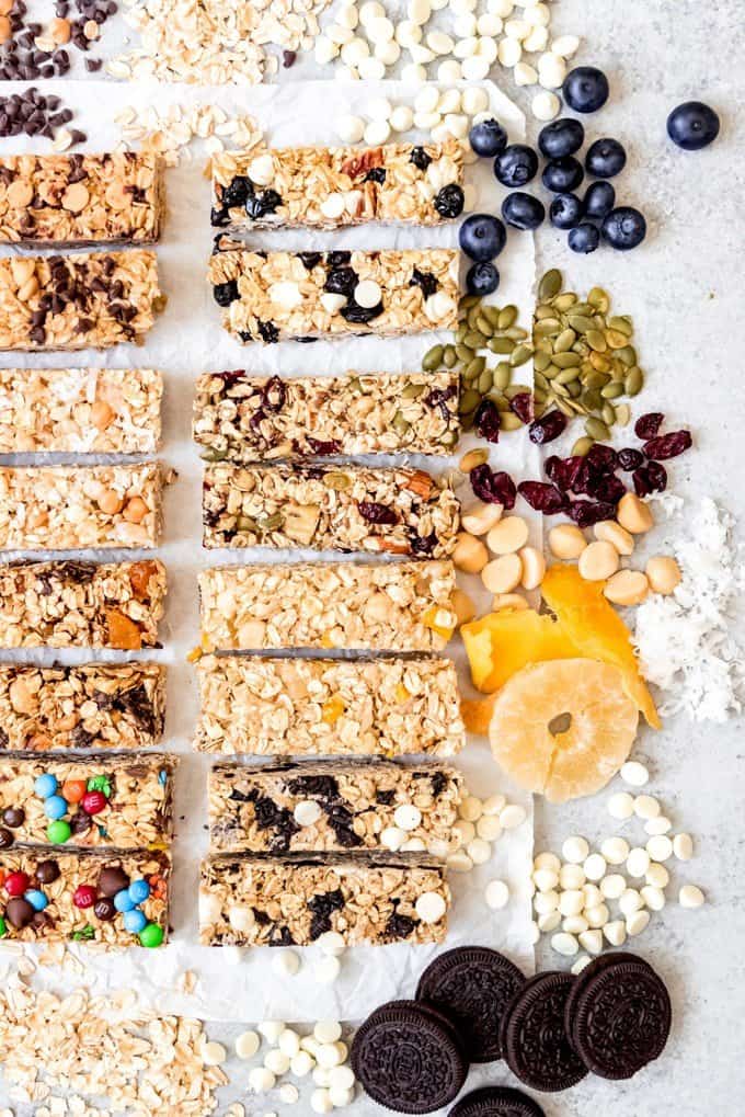 An image of chewy granola bars arranged next to some of the ingredients used to make them in different flavors like dried pineapple, dried mango, dried cranberries, pumpkin seeds, Oreos, and white chocolate chips.