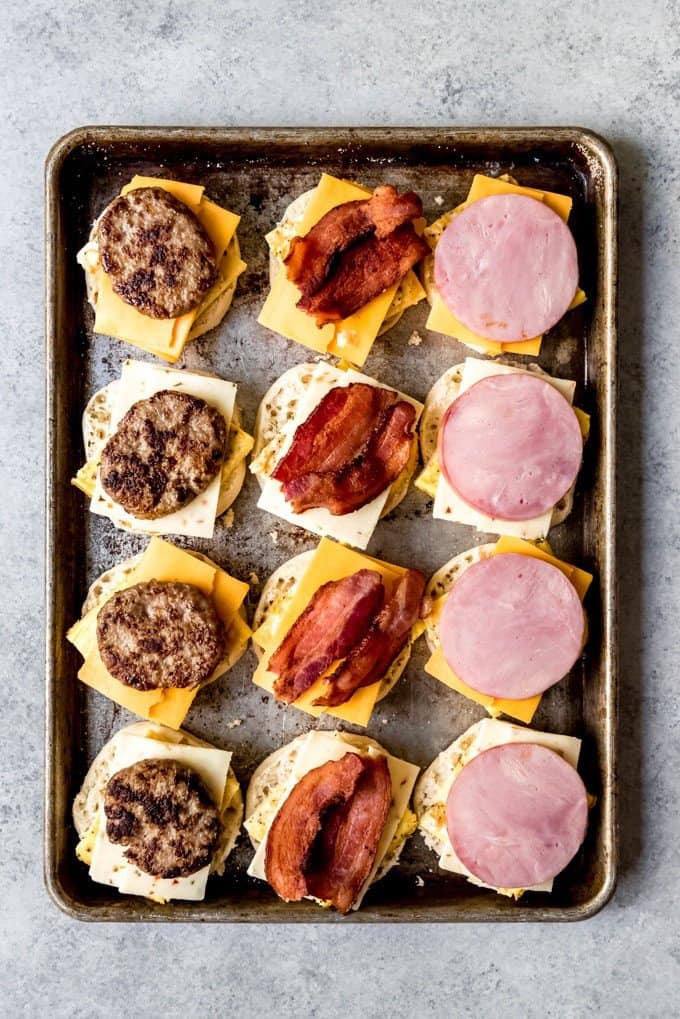 An image of freezer breakfast sandwiches being assembled with sausage, bacon, and ham over baked eggs, cheese, and english muffins.