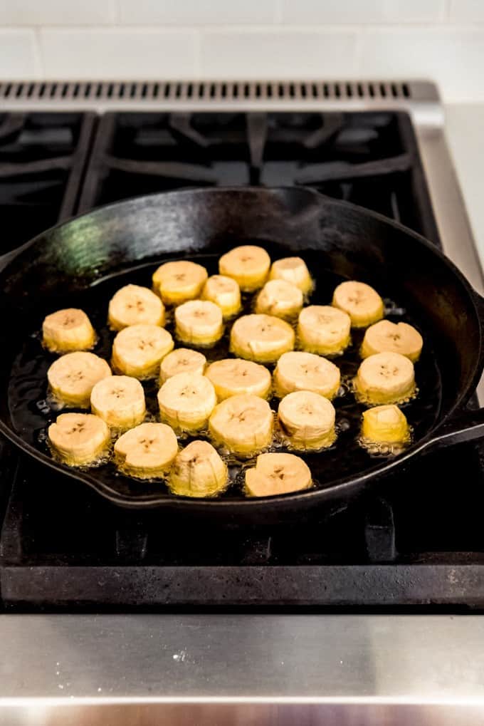 An image of plantains frying in oil in a cast iron skillet.