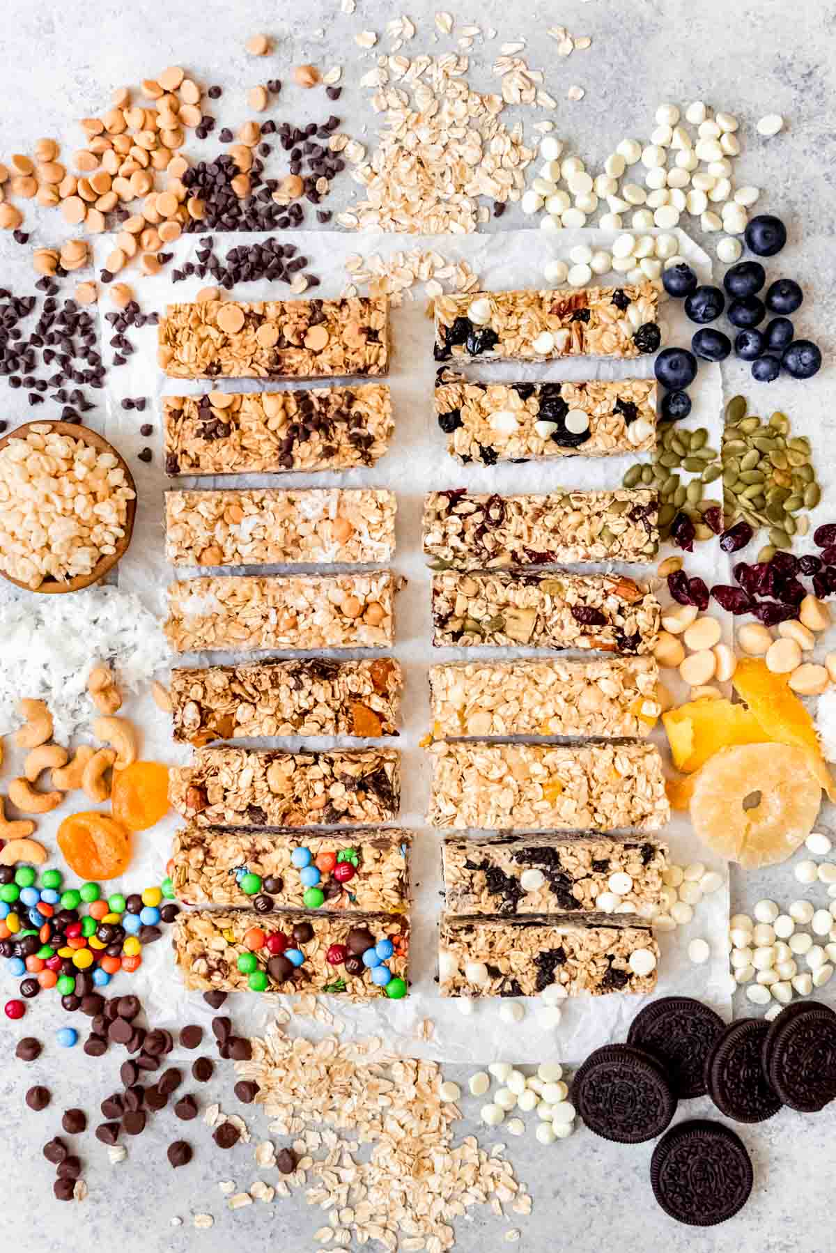 An image of eight different flavors of homemade granola bars arranged on a surface with ingredients spilling around them.