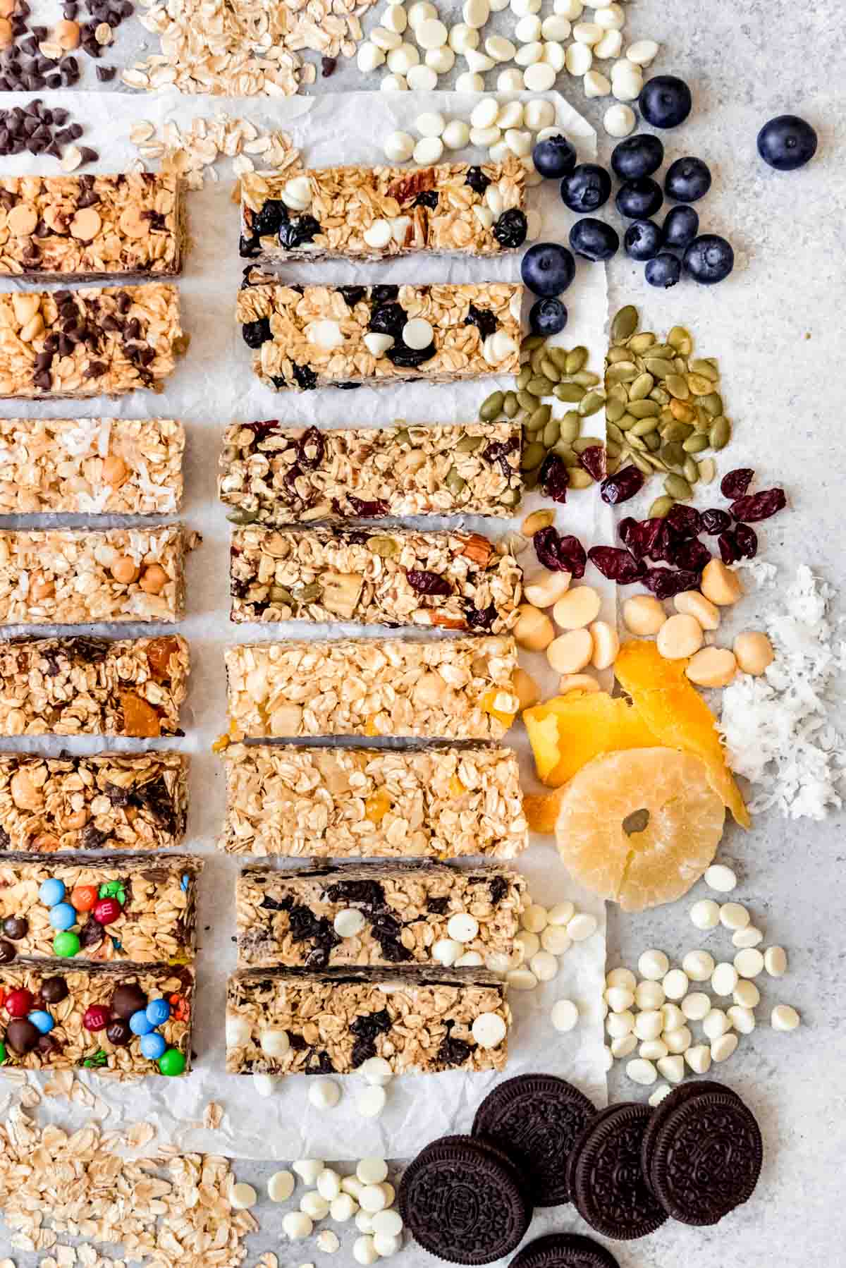 An image of chewy granola bars arranged next to some of the ingredients used to make them in different flavors like dried pineapple, dried mango, dried cranberries, pumpkin seeds, Oreos, and white chocolate chips.