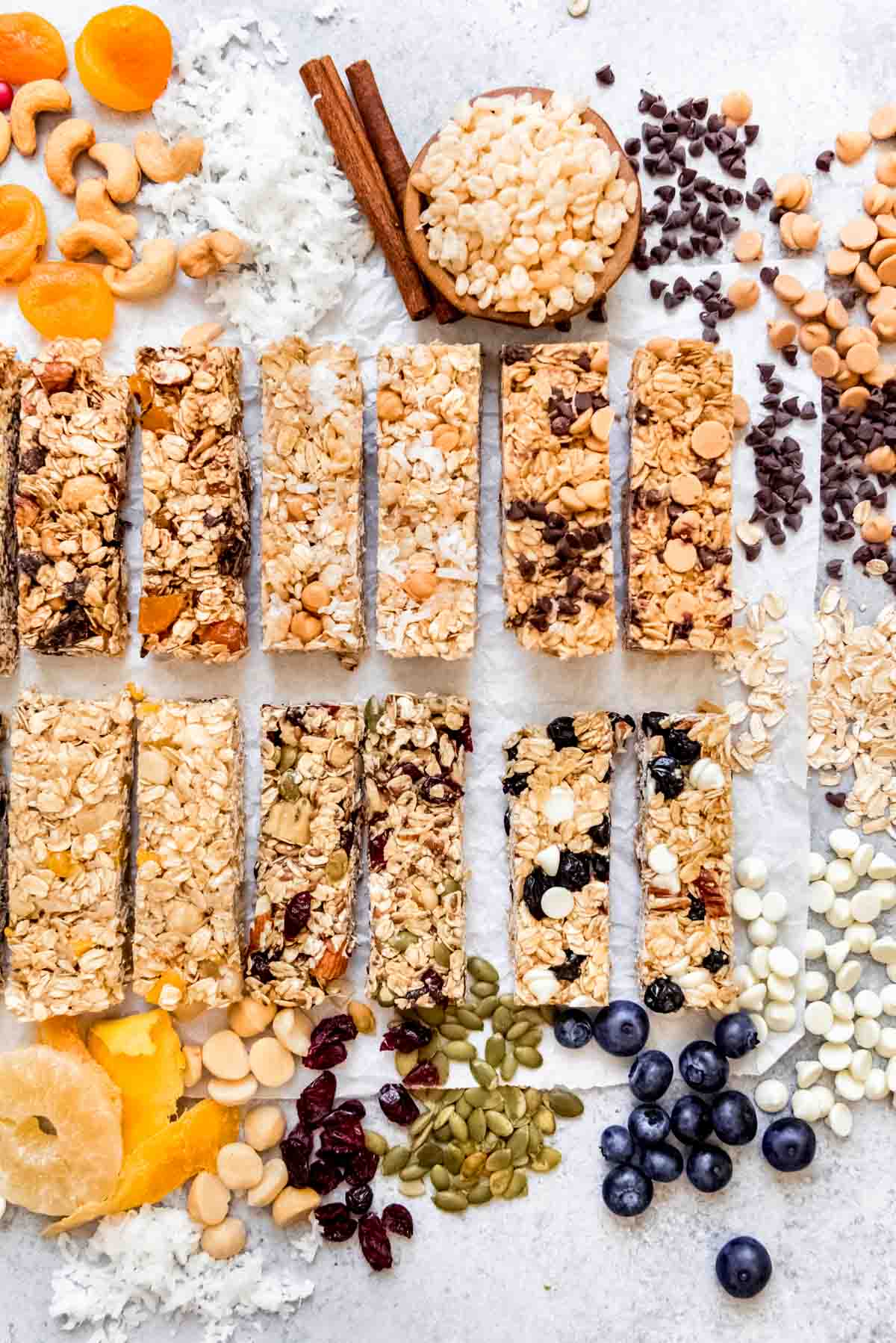 An image of six different varieties of homemade granola bars arranged two-by-two on a surface with some of the ingredients around them to show the flavors.