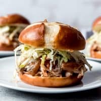 An image of a pulled chicken sandwich made in the slow cooker with a pretzel bun, apple barbecue sauce, and an apple cabbage slaw.