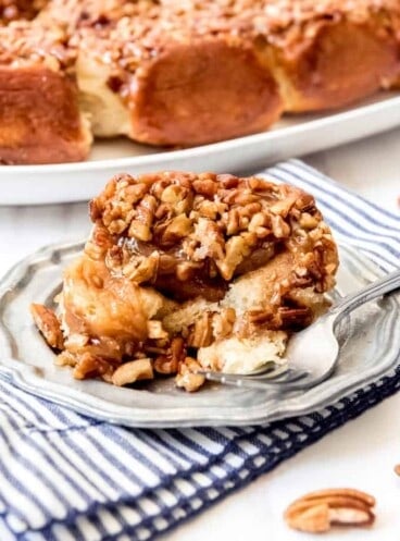 A plate with a caramel pecan sticky bun and a fork while resting on top of a blue and white striped towel in front of a plate full of caramel pecan sticky buns