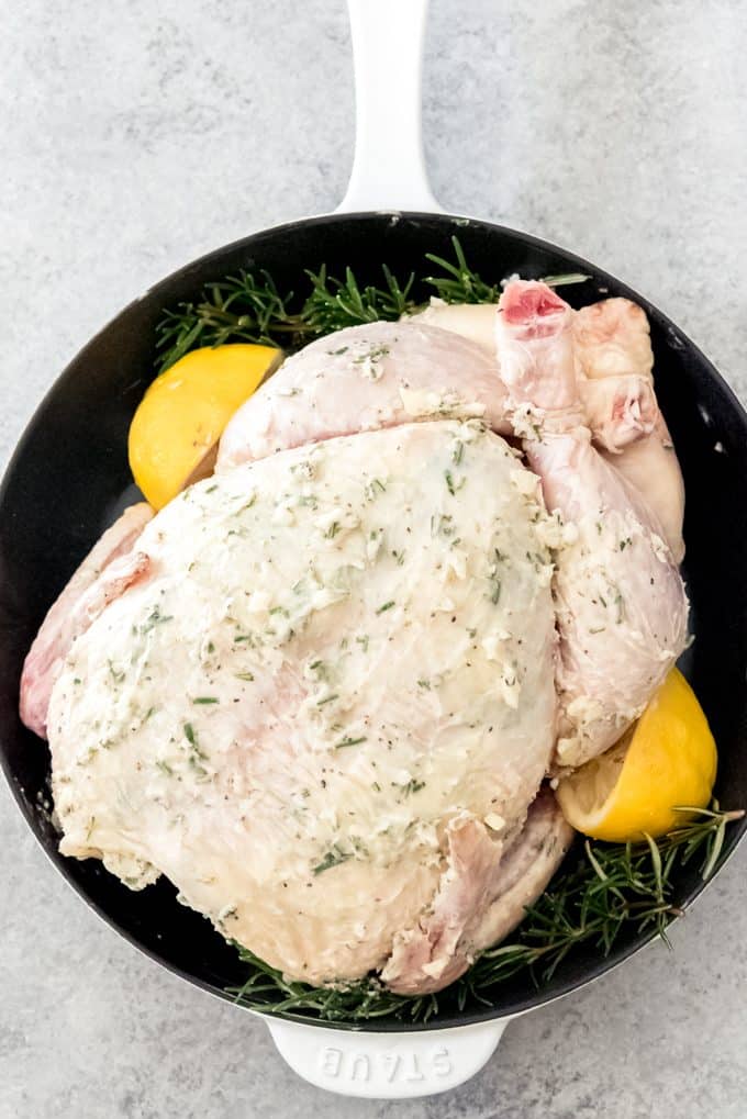 whole chicken in roasting pan (dutch oven) covered in garlic butter, beside rosemary and lemon
