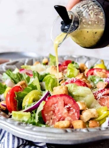 An image of homemade Olive Garden salad dressing being poured over a big bowl of salad with lettuce, tomatoes, red onions, croutons, and pepperoncini peppers.