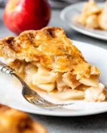 An image of a slice of homemade apple pie on a white plate with a red apple behind it.