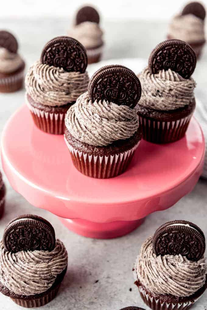 An image of Oreo frosting piped in swirls onto moist chocolate cupcakes sitting on a pink cake stand.