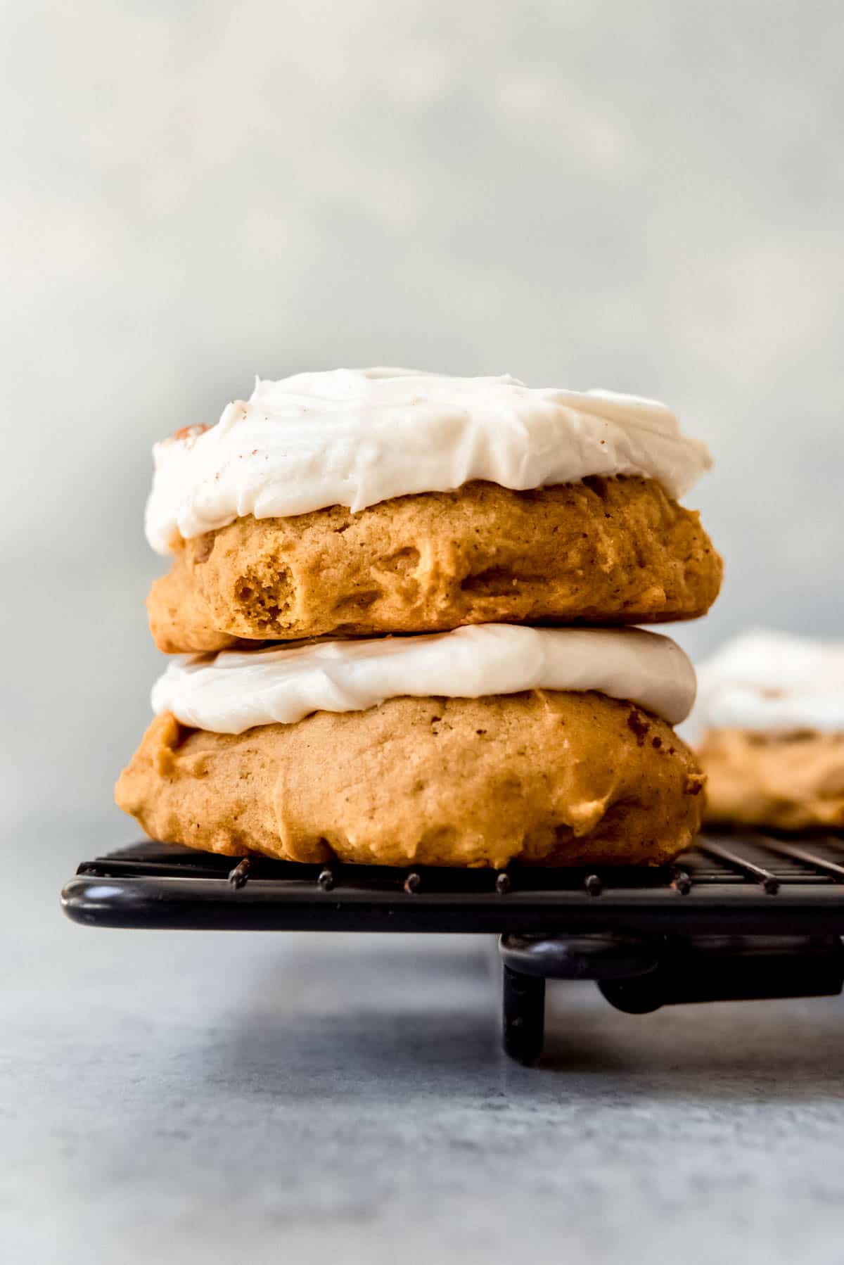 An image of two pumpkin cookies with cream cheese frosting stacked on top of each other on a black wire cooling rack.