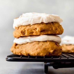 An image of two pumpkin cookies with cream cheese frosting stacked on top of each other on a black wire cooling rack.