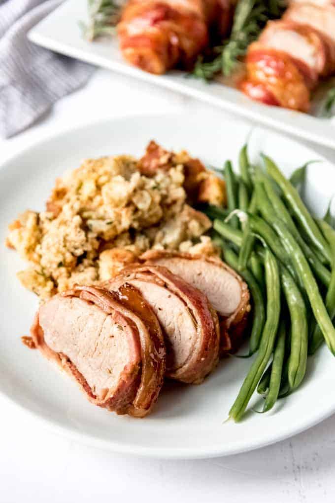 Another image of a bacon-wrapped pork tenderloin on a white plate with cornbread stuffing and green beans.
