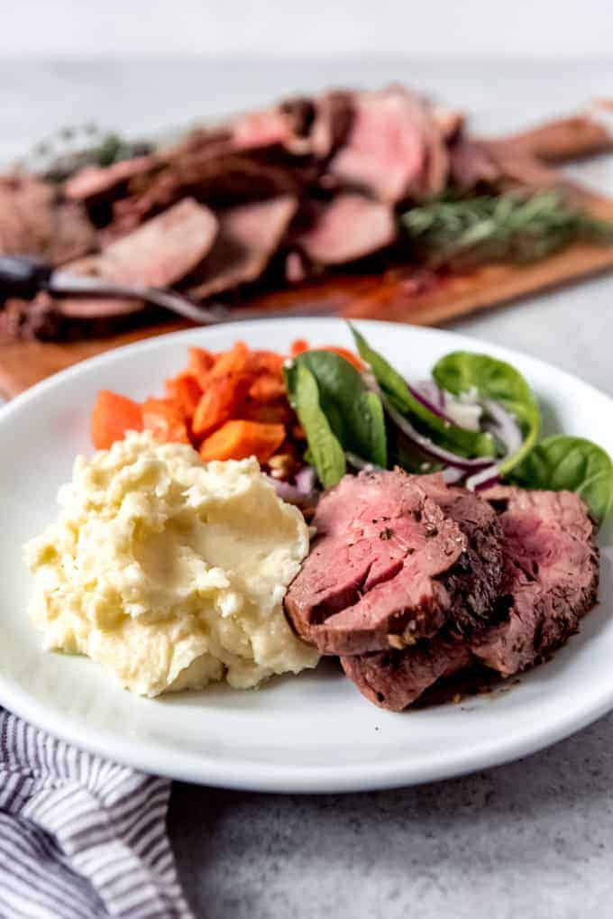An image of a plate with garlic herb butter roasted beef tenderloin, creamy mashed potatoes, salad, and roasted carrots.