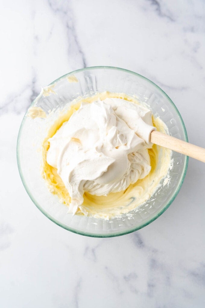 Adding whipped cream to a cream cheese and white chocolate mixture in a glass mixing bowl.