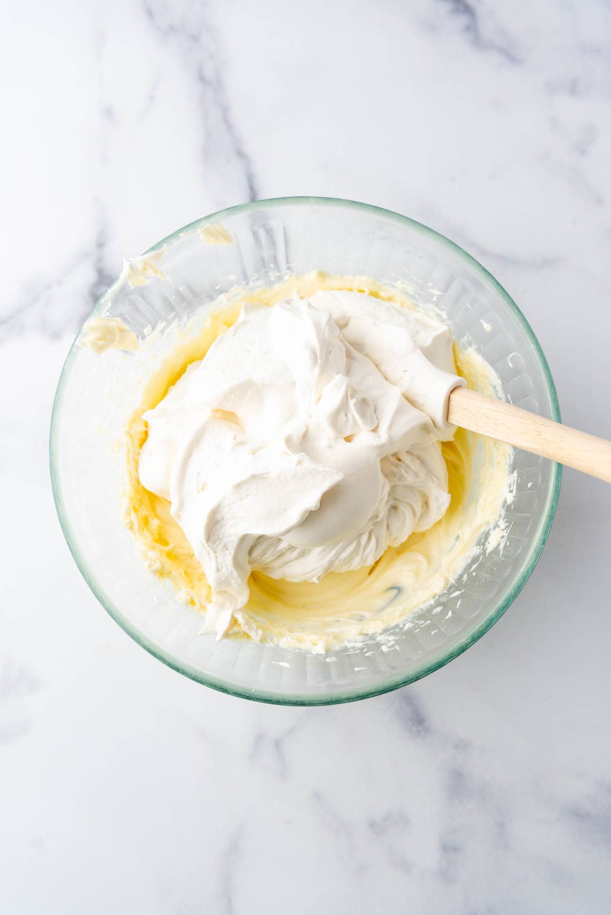 Adding whipped cream to a cream cheese and white chocolate mixture in a glass mixing bowl.