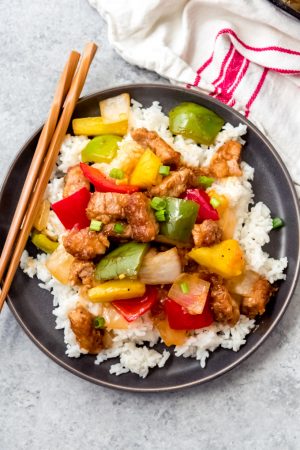 a gray plate topped with a pile of rice and covered in sweet and sour pork with wooden chopsticks to the side