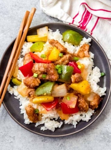 a gray plate topped with a pile of rice and covered in sweet and sour pork with wooden chopsticks to the side