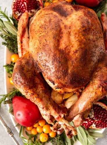 An image of a nicely browned whole Thanksgiving turkey on a serving platter surrounded by fruit and herbs.