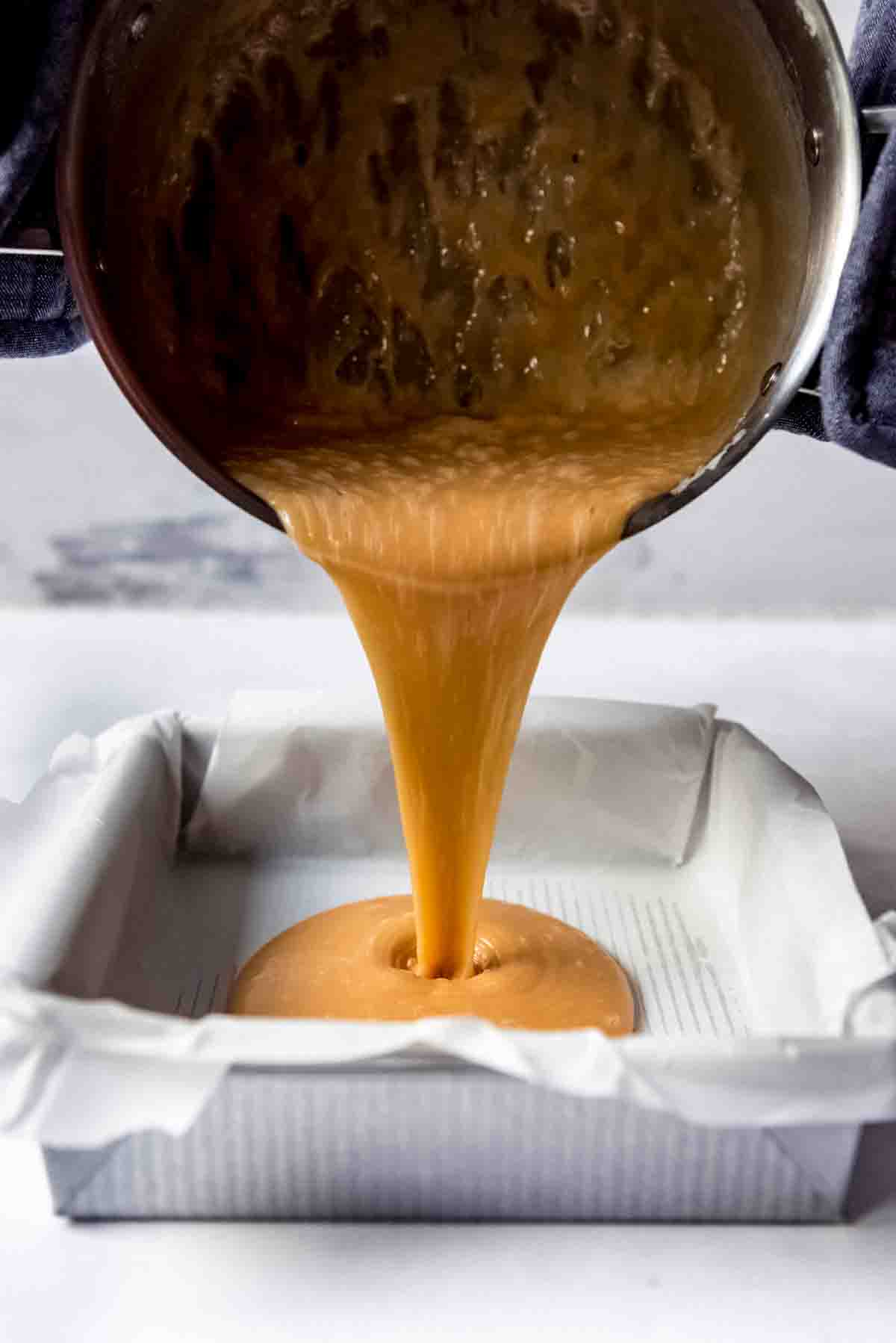 Caramel being poured from a pot into a square baking pan lined with parchment paper.